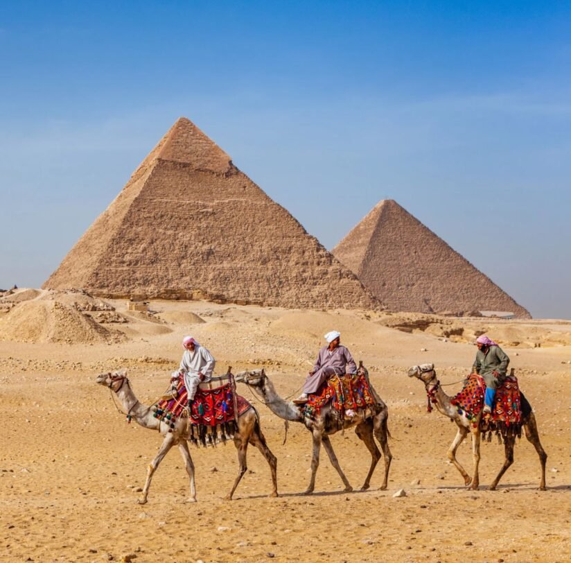 Three People Riding Camels In Front Of The Great Pyramids Of Giza Under A Clear Blue Sky